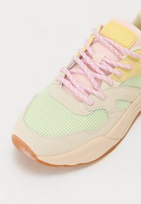 Sneaker leggera con pannelli in rete verde menta e suede beige, lacci a fantasia rosa e una suola in gomma testurizzata. Etichetta del logo visibile.
