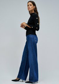 Suéter negro ajustado con detalles en botones dorados en las mangas, combinado con jeans de mezclilla azul de pierna ancha y tacones negros de punta. 