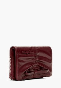 Cartera de cuero burdeos con un detalle de lazo en el frente, cremallera en el lateral y logo "Desigual" grabado.