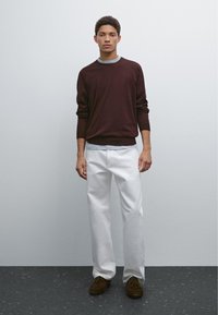 Maglione bordeaux con rifiniture grigie, abbinato a pantaloni bianchi a gamba dritta. Il modello indossa scarpe marroni scure. Design semplice con uno sfondo neutro.