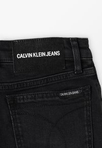 Vaqueros de mezclilla negra con una etiqueta tejida que dice "CALVIN KLEIN JEANS." Presenta costuras visibles y una textura suave. Detalle distintivo en el bolsillo trasero.