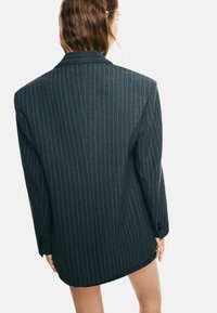 Donna con capelli alla spalla che indossa un blazer oversize a righe scure, girata di spalle contro uno sfondo chiaro e semplice.