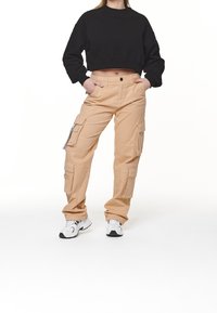 Beige Cargo-Hosen mit mehreren Taschen, Knopfverschluss und lässigem Schnitt, kombiniert mit einem schwarzen cropped Sweatshirt und weißen Turnschuhen.