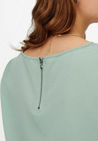 Blouse vert clair avec une texture lisse, dotée d'une courte fermeture éclair au dos et d'un col rond. Détails de surpiqûre subtils le long des bords.