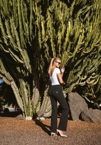 Mustat leveälahkeiset farkut, joissa on takataskujen yksityiskohta, yhdistetty valkoiseen istuvaan toppiin ja ihonvärisiin korkokenkiin, suurta vihreää kaktusta vasten.