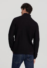 Uomo con capelli ricci che indossa un maglione a collo alto nero texture e jeans blu scuri, visto di spalle su uno sfondo semplice.