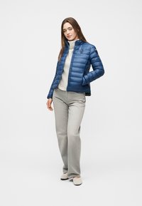 Γυναίκα που φοράει μπλε μπουφάν τύπου puffer πάνω από λευκό πουλόβερ με ρολό λαιμό, γκρι παντελόνι φαρδύ στο τελείωμα και μπεζ παπούτσια, στέκεται με το ένα χέρι στην τσέπη.