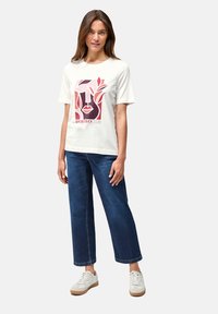 Camiseta blanca de algodón con un gráfico colorido que presenta un rostro estilizado y follaje. Combinada con unos vaqueros de pierna ancha azules y zapatillas blancas.