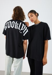 Svarta grafiska t-shirts har fet vit "Brooklyn" text i stiliserad font på ryggen, med avslappnad passform och mjukt bomullsmaterial.