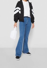 Chaqueta bomber negra con rayas blancas, camiseta polo blanca, jeans azules de campana y zapatillas negras. Sosteniendo una bolsa blanca con cremallera.