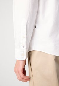 De linkerhand van een man rust nabij de zak van een beige broek, terwijl hij een wit overhemd met lange mouwen draagt met een knoopmanchet en kleine multikleurige draaddetails.