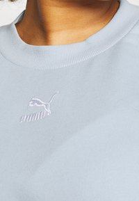 Lähikuva vaaleansinisestä Puma-hupparista, jossa on brodeerattu Puma-logo kankaassa ja osittainen näkymä kaulasta ja leuasta.
