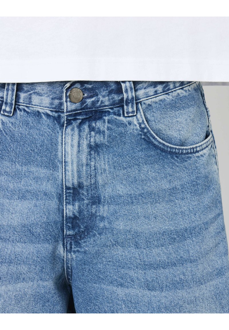 Jeans en denim bleu avec un lavage délavé, design à cinq poches, fermeture à bouton en métal et coupe droite. La texture semble lisse avec de légères variations.