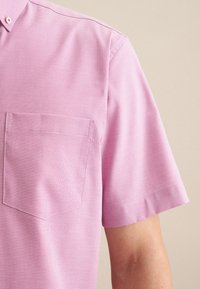 Camicia rosa a maniche corte con tessuto testurizzato, dotata di taschino sul petto e colletto con bottoni, che mostra un design semplice con dettagli discreti.