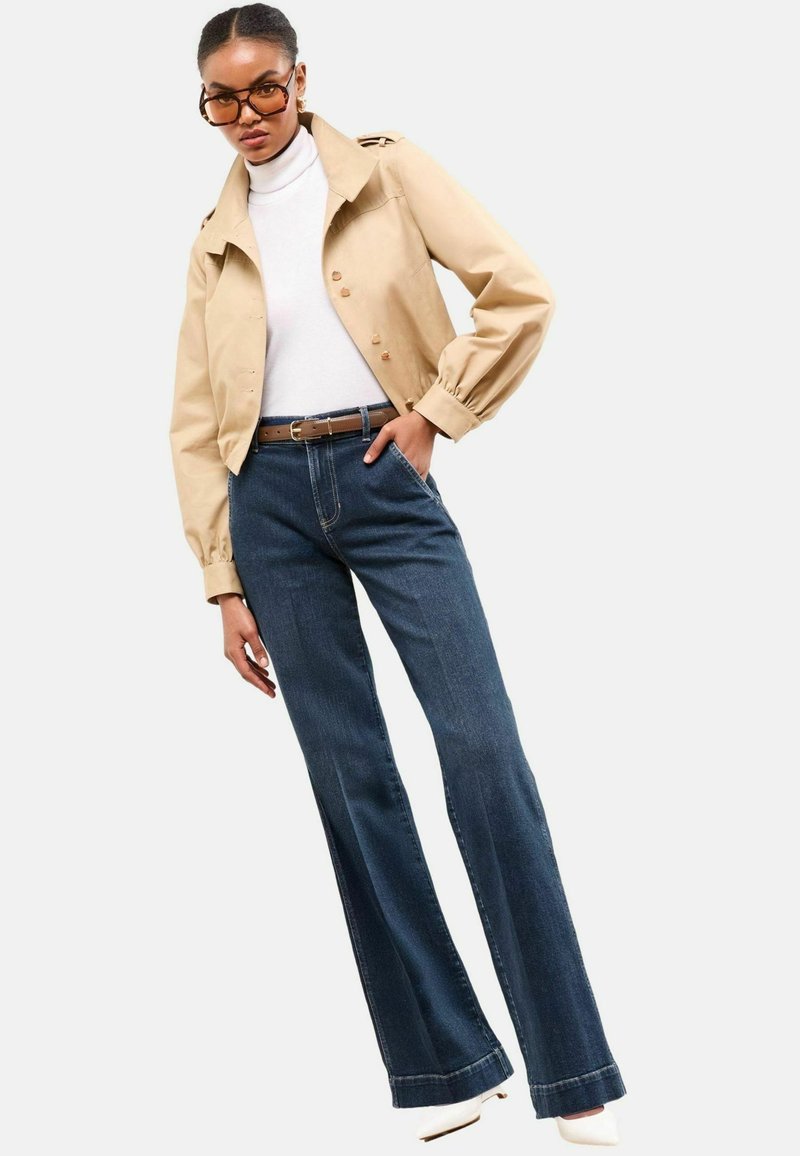 Beige cropped jack met pofmouwen, witte coltrui, donkerblauwe flare jeans en witte hakken. Overmatige bruine zonnebril.