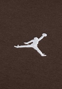 Material maro cu o suprafață texturată, având un logo Jumpman brodat alb, care înfățișează o figură într-o poziție de zbor ținând o minge de baschet.