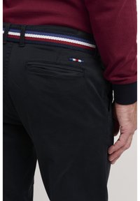 Black pants with a textured finish, featuring a multicolor woven belt with red, white, and blue stripes. A small tri-colored accent on the pocket.