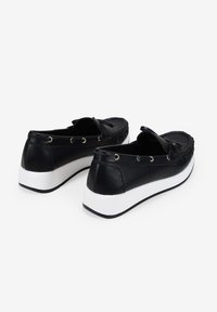 Zapatos negros de piel sin cordones con una suela plana blanca, que presentan costuras decorativas y ojales metálicos a lo largo de los costados.