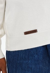 Witte gebreide trui met een bruin "blue shadow" label, gedragen over blauwe denim jeans, met een zichtbare arm van een donkere huidskleur.