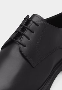 Μαύρο δερμάτινο επίσημο παπούτσι με κλειστό κορδόνι και τακτοποιημένες ραφές, εμφανίζεται σε κοντινό πλάνο σε ουδέτερο φόντο.