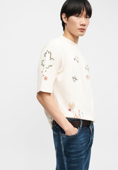 Νεαρός άνδρας με μαύρα μαλλιά που φοράει κρεμ κοντομάνικο πουκάμισο με πολύχρωμα κεντημένα και διακοσμημένα σχέδια, μπλε τζιν, χέρι στην τσέπη, κοιτάζοντας στο πλάι.