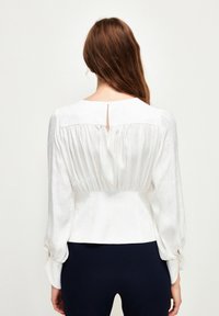 Witte blouse met een geplooide achterkant, lange mouwen, splitnek en een zachte textuur. Details met elastische manchetten en een aansluitende taille.