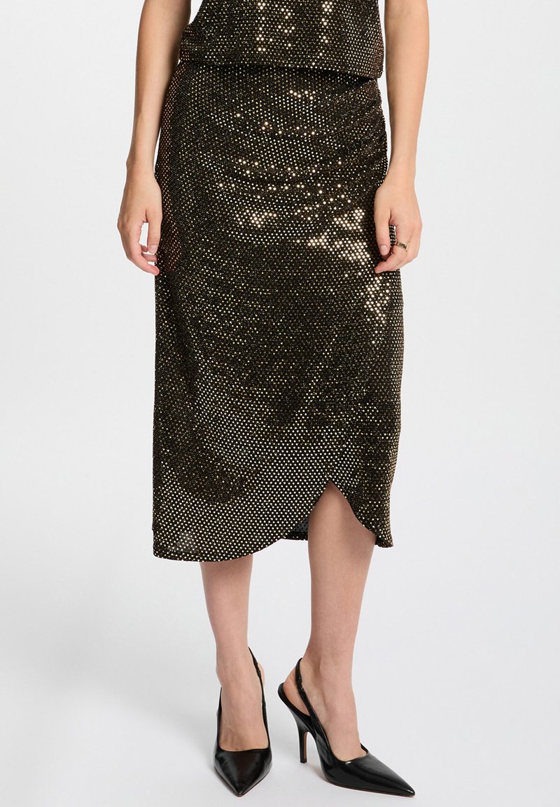 Femme portant une jupe midi noire ornée de sequins dorés et d'une fente sur le côté, associée à des escarpins à talons aiguilles noirs à brides arrière.