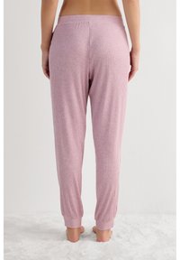 Vrouw staat op blote voeten op een wit, pluizig tapijt, gekleed in lichtroze, geribbelde loungebroek, gezien van achteren.