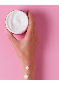 Witte crème in een ronde rode pot die in een hand wordt vastgehouden, met twee dotjes crème op een pols tegen een roze achtergrond.