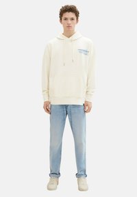 Felpa crema con tasca frontale e cappuccio con cordino, con branding blu sul lato sinistro del petto, abbinata a jeans blu chiaro e sneakers crema.