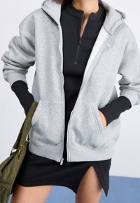 Donna che indossa una felpa grigia con zip sopra un vestito nero con spacco frontale, con una borsa in velluto a coste verde oliva sulla spalla.