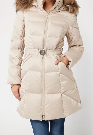 Femme portant un manteau d'hiver matelassé beige avec une capuche bordée de fourrure, une taille ceinturée et une fermeture éclair à l'avant, les mains dans les poches, associée à un jean bleu.