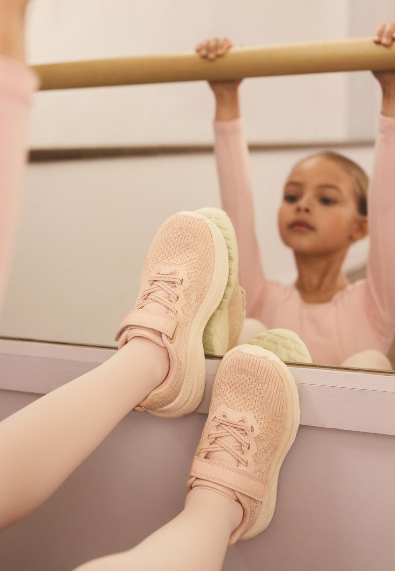 Scarpe sportive rosa chiaro con tomaia in maglia strutturata, chiusura con lacci e velcro, suola in gomma bianco-verde, mostrate su uno sfondo riflettente.