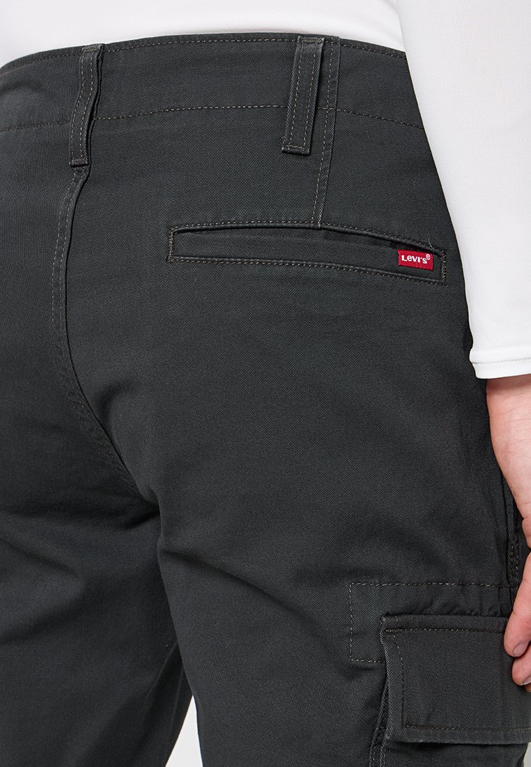 Pantalones negros de Levi's con bolsillos traseros y laterales, llevados con una camisa blanca de manga larga, mostrando una mano descansando cerca del bolsillo lateral.