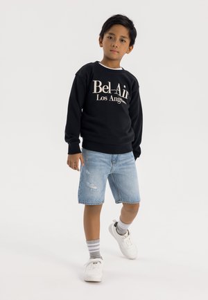 Melna kapuce ar "Bel-Air Los Angeles" beigu tekstā, kopā ar nodilumu redzošiem džinsu šortiem un baltām kedām. Redzami pelēki svītraini zeķes.