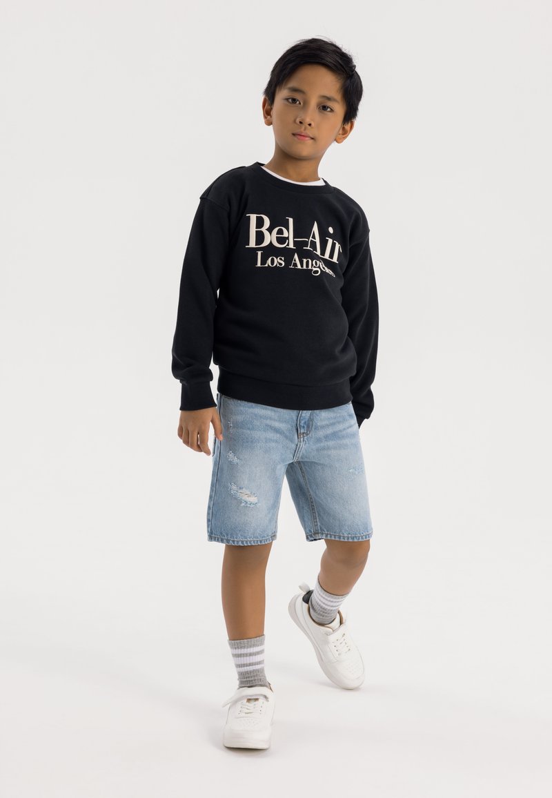 Sudadera negra con "Bel-Air Los Ángeles" en texto beige, combinada con unos pantalones cortos de mezclilla desgastados y zapatillas blancas. Medias gris a rayas visibles.