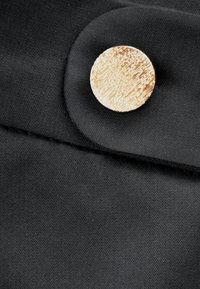 Czarna tkanina o gładkiej fakturze z okrągłym, teksturowanym złotym guzikiem, umieszczonym na złożonym fragmencie. Wyraźna linia szwu nad guzikiem.