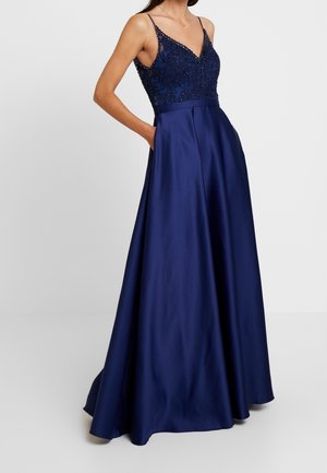Robe de soirée bleu marine sans manches avec corsage en dentelle perlé, encolure en V et jupe en satin pleine, portée avec une main dans la poche.