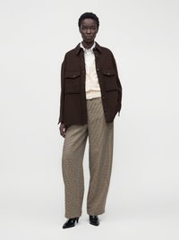 Femme debout portant une veste oversize marron foncé, un pull crème sur une chemise blanche, un pantalon large à carreaux beige et des chaussures noires pointues.