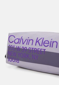 Torba Calvin Klein z jasnofioletowego materiału, z wyraźnym, kontrastującym fioletowym napisem zawierającym nazwę marki i adres w nowoczesnej czcionce.