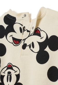Chemise en coton crème avec un imprimé Mickey Mouse noir répété et des pois. Présente une fermeture à pression au niveau du col.