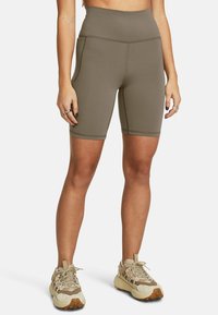 Athlète portant un short de vélo taille haute vert olive et des baskets beiges à semelles épaisses, se tenant avec les bras détendus le long du corps.