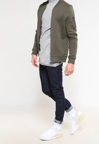 Olive green bomber jacket over a grey sweatshirt, paired with dark denim jeans and white sneakers with light accents. Features zippered pocket.