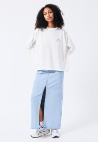 Sudadera de gran tamaño en color blanco roto con un gráfico azul, combinada con una falda maxi de mezclilla azul clara que presenta una abertura frontal. Completada con zapatillas blancas.