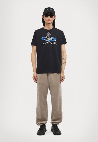 Black cotton T-shirt with "ANTI-WASTE" graphic, paired with beige velour pants and black sneakers. Model wearing sunglasses and a cap.