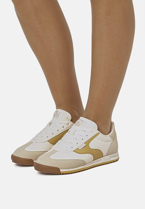 Zapatillas blancas y beige con suelas marrones y detalles amarillos en los costados, usadas en pies descalzos sobre un fondo liso.