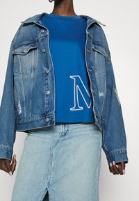 Chaqueta de mezclilla con botones de cobre y detalles desgastados, superpuesta a una camiseta azul con un gráfico blanco de la letra "N", usada con una falda azul clara.
