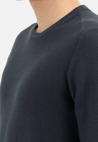 Pull en maille bleu foncé avec un col rond, un tissu texturé et des détails côtelés aux épaules et au col.