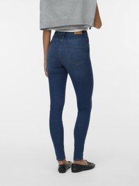 Jeans de ganga azul skinny com bolsos traseiros, uma cintura alta e detalhes de costura mínimos. Combinados com sapatos pretos planos e uma blusa cinza.