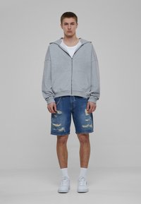 Sudadera gris con cremallera, camiseta blanca debajo, pantalones cortos de mezclilla desgastados, zapatillas blancas, y cordones a la altura de la rodilla, con un acento de cadena en los pantalones cortos.